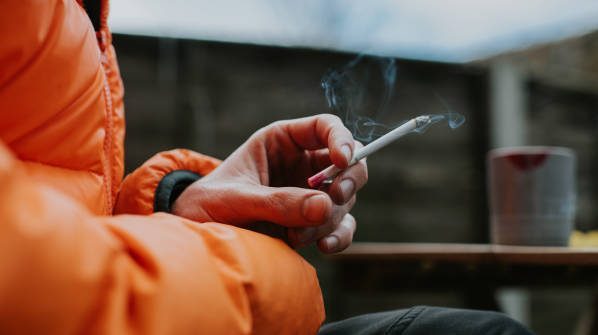 A man sits outside and holds a slim menthol cigarette between his fingers. Close-up with focus on the cigarette.