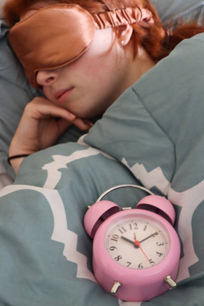 Stock photo showing close-up view of a pink metal case retro alarm clock with double bell on duvet. An attractive, redheaded woman can be seen lying in the double bed, sleeping with an eye mask.